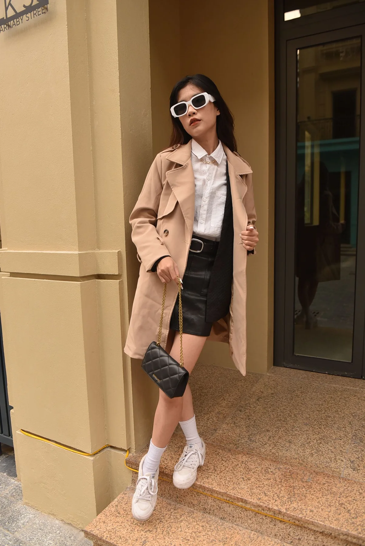 Woman in a beige coat and sunglasses standing on a city street.